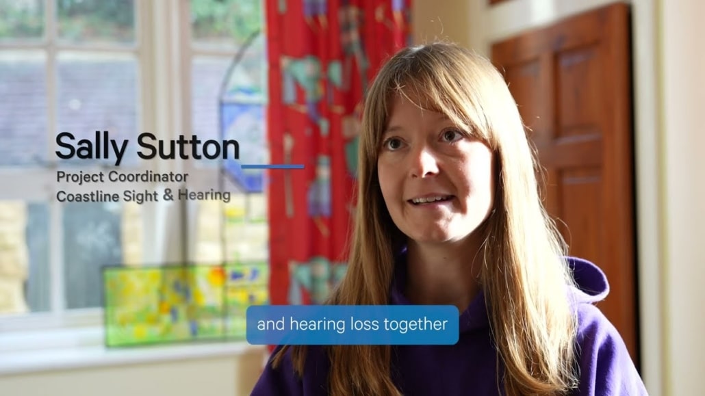 A woman with long light brown hair, wearing a purple hoodie, sits indoors in a well-lit room. Text on the image reads: “Sally Sutton, Project Co-ordinator, Coastline Sight & Hearing” and “and hearing loss together.”.
